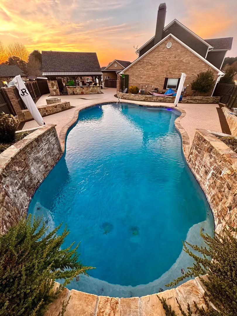 Sunset view of a pool with stone knee walls and tan pebble-finish deck surfacing