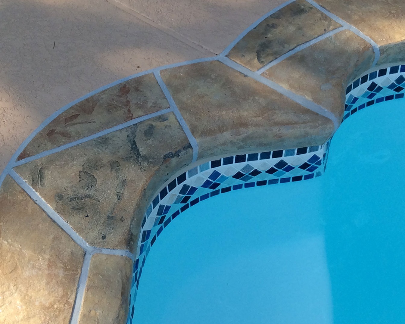 Close-up of tan stone coping and blue mosaic tile band framing a pool corner
