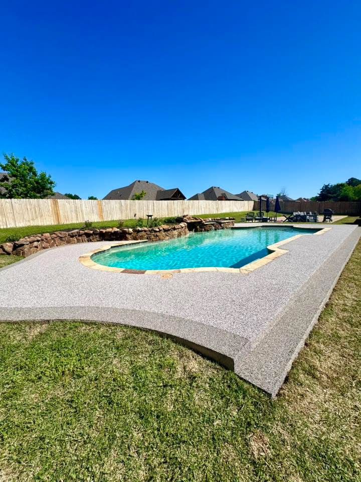 Freeform pool with gray speckled deck surrounded by a wood fence and grass yard